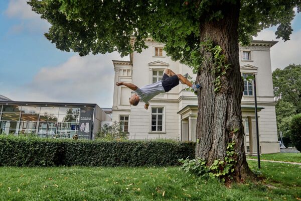 Junger Mann macht einen Backflip an Baum vor dem Landesmuseum